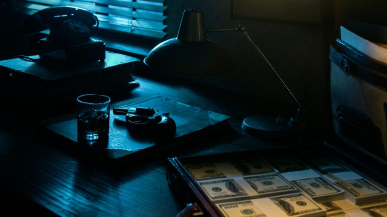 Vintage film noir style desktop with revolver, drink and open briefcase full of cash money: mafia, crime and thriller concept